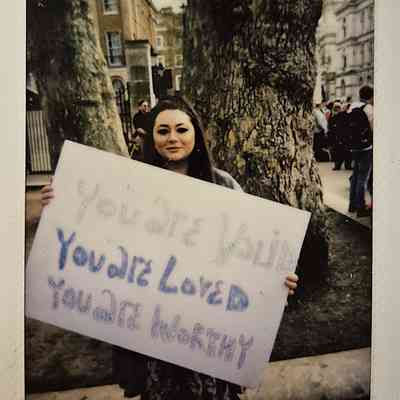 A woman stands outdoors holding a sign that reads, "You are valid, you are loved, you are worthy".
