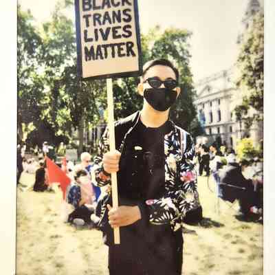 A person holding a "Black Trans Lives Matter" sign stands outdoors, wearing sunglasses and a mask.