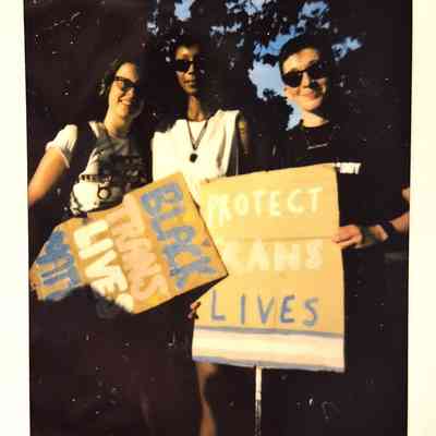 Three individuals are holding signs advocating for Black trans lives, standing outdoors under a tree.