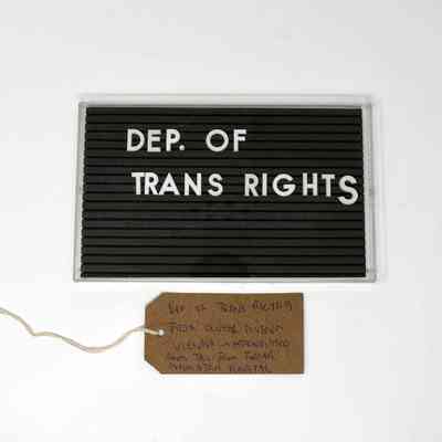 A black Marquee style sign board with the lettering "DEP. OF TRANS RIGHTS" in white letters. With a brown, handwritten luggage tag.