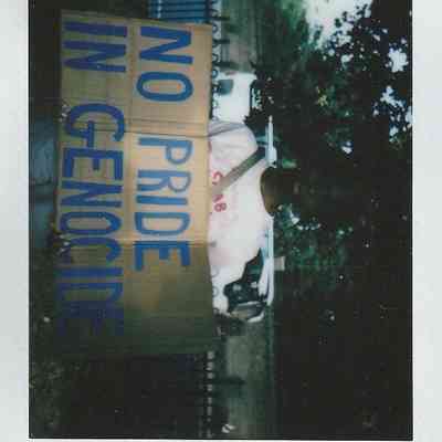 A person holds a cardboard sign with "No Pride in Genocide" written in bold blue letters.