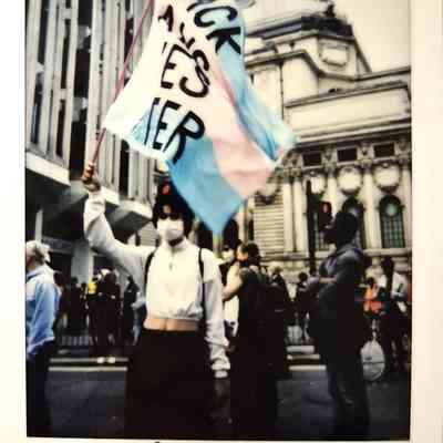 A person wearing a mask holds a trans flag during a protest in London, with buildings visible behind.