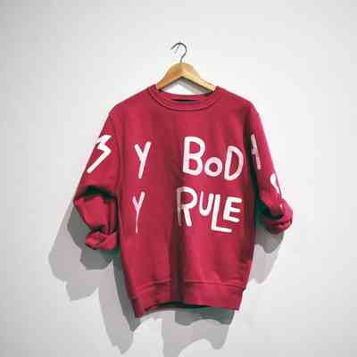 A red sweatshirt with white text hangs on a wooden hanger against a plain wall.