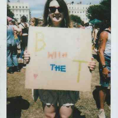 A person at an outdoor event holds a sign reading "B with the T" while smiling.