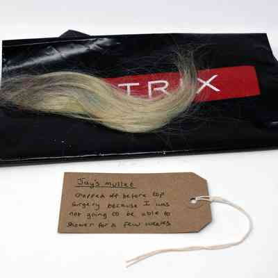A section of blonde hair lays on a black plastic bag with the text "MATRIX" in white on a red background. With a brown, handwritten luggage tag.