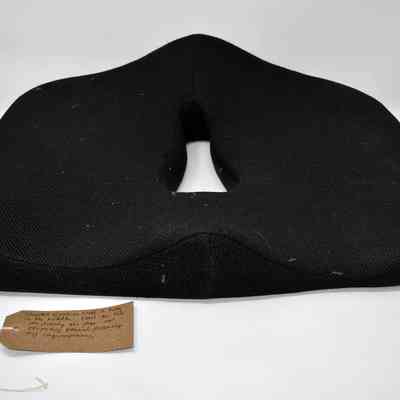 A black, shaped donut cushion for use after vaginoplasty surgery. With a brown, handwritten luggage tag.