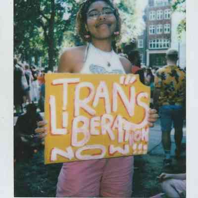 A person holds a vibrant sign reading "Trans Liberation Now" at an outdoor gathering under trees.