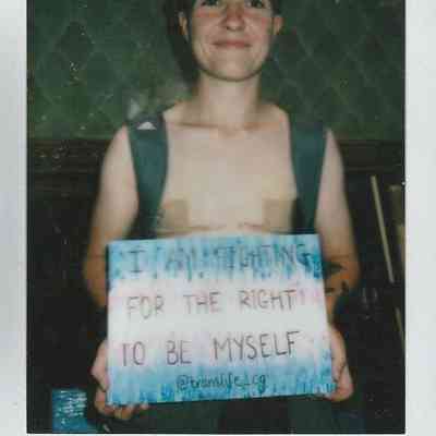 A person smiles while holding a sign advocating for the right to self-identity and expression.
