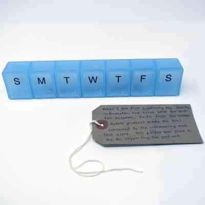 A transparent blue pill box that has seven compartments, labeled for the days of the week. With a brown, handwritten luggage tag.