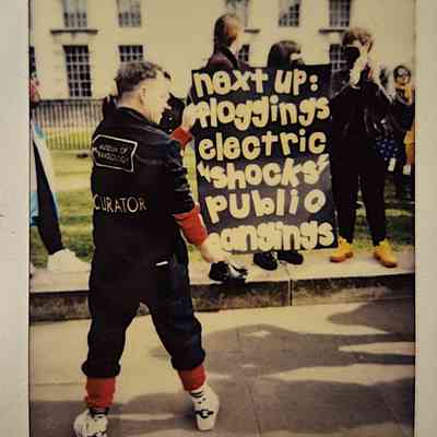 A person stands in front of individuals holding a handmade sign which says "NEXT UP: FLOGGINGS ELECTRIC SHOCKS PUBLIC HANGINGS".