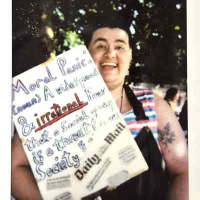 A person holds a colorful sign about moral panic, smiling in a sunny outdoor setting.