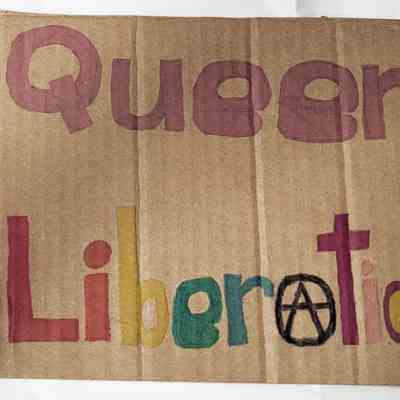 The image features a cardboard sign stating "Queer Liberation".