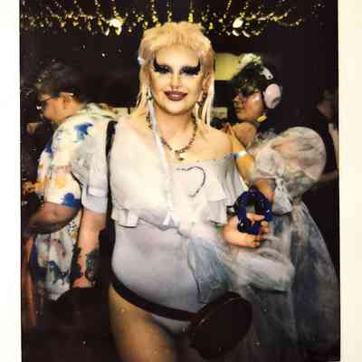 A person in elaborate makeup, wearing a stylish outfit, holds a tambourine in a festive space.