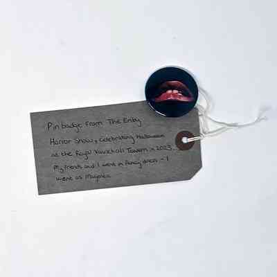 A small badge featuring red lips on a black background. The image is synonymous with the Rocky Horror Picture Show. With a brown, handwritten luggage tag.