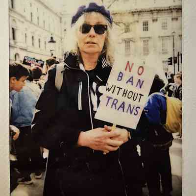 A person wearing sunglasses holds a sign reading, "NO BAN WITHOUT TRANS," at a protest.