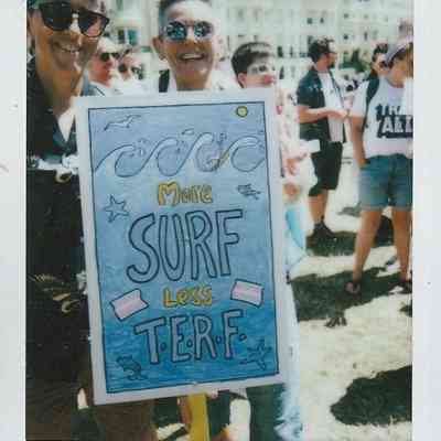 People are smiling outdoors, holding a sign reading "More Surf Less TERF" during a sunny day.