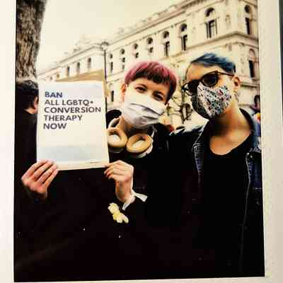 Two people wearing masks hold a sign that reads, "Ban all LGBTQ+ conversion therapy now" outside.