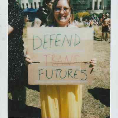 A person in glasses holds a sign reading "Defend Trans Futures" at an outdoor event.