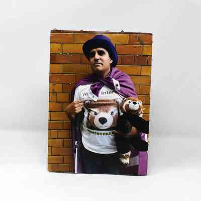 The person is wearing a purple flag and hat, holding a stuffed animal, standing against a tiled wall.