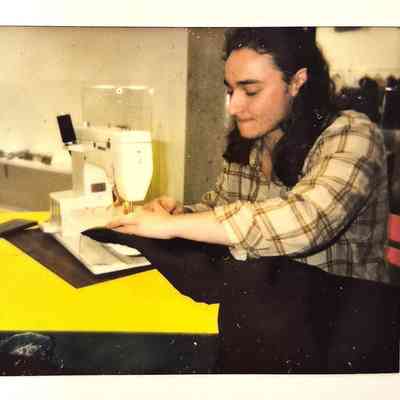 A person wearing a plaid shirt is sewing fabric on a white sewing machine indoors.