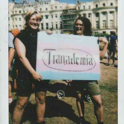 Two people standing outdoors hold a colorful sign labeled "Tranademia".