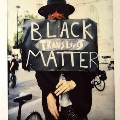 A person wearing a hat and mask holds a sign reading "Black Trans Lives Matter" at a protest.