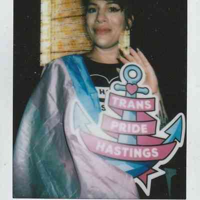 A person proudly holds a "Trans Pride Hastings" sign while wearing a flag at an event.