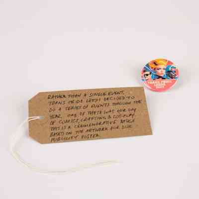 A small colourful badge with text "TRANS PRIDE LEEDS 2023" with a background of three stylised faces. With a brown, handwritten luggage tag.