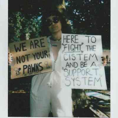 A person holds two protest signs while wearing sunglasses outdoors in a park setting with trees.