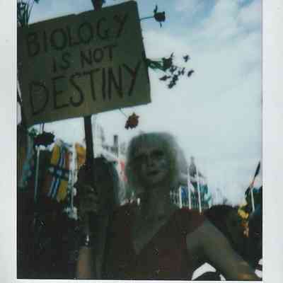 A person with blonde hair holds a protest sign that reads "Biology is not destiny".