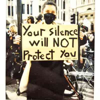 A masked protester holds a sign reading, "Your silence will NOT protect you," at a demonstration