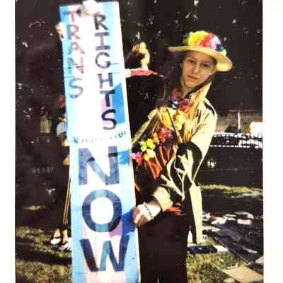 A person in a colorful hat holds a sign reading "Trans Rights Now" during an outdoor gathering.