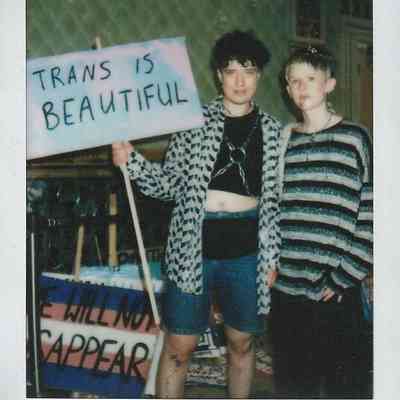Two individuals stand together, one holding a sign that reads "Trans is Beautiful," expressing support and pride.