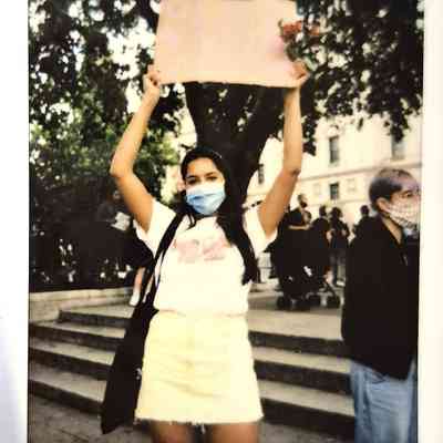 A person wearing a mask holds a sign overhead at a protest, standing on steps.