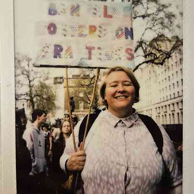 A smiling person holds a protest sign outdoors, surrounded by people and trees in the background.