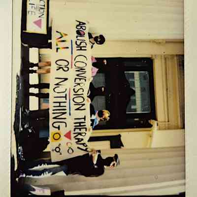 A group stands on steps holding a banner reading "Abolish conversion therapy, all or nothing."