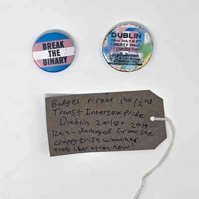 Tag reading "Badges from 1st/2nd Trans + Intersex Pride Dublin 2018 + 2019
Rain-damaged from the crappy Irish weather.
Trans liberation now!" Under the tag are two badges. Badge 1 reads "DUBLIN 28th July @ 2PM LIBERTY HALL TO FAIRVIEW PARK" and has a trans flag design background. Badge 2 reads "BREAK THE BINARY" and has a trans flag background.