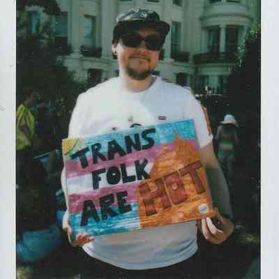 A person wearing sunglasses holds a colorful sign reading "Trans folk are hot" outside a large building.