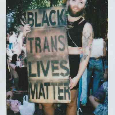 A person stands outside holding a large sign that reads "Black Trans Lives Matter" at a gathering.
