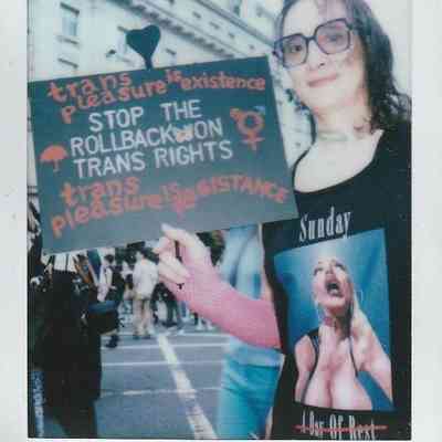 A person holds a sign which says "Trans Pleasure Is Existence Stop The Rollback On Trans Rights Trans Pleasure Is Resistence".