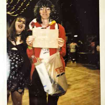 Two people smile cheerfully in a festive setting, with one holding a sign and bags.