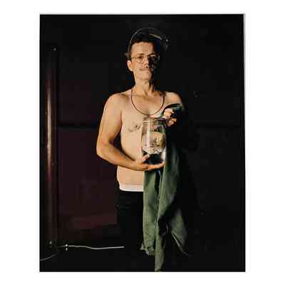 Colour photograph mounted onto metal back, unframed & unglazed; matt finish image of E-J Scott wearing green camouflage baseball cap, metal framed glasses, bare to waist, black trousers. Holding Kilner jar & green shirt. Jar contains human chest tissue. Image taken against dark background.