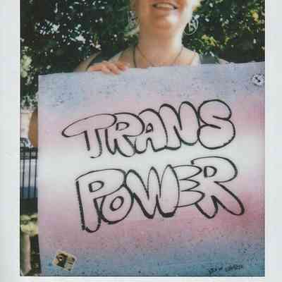 A person wearing sunglasses and a hat holds a sign reading "Trans Power" with trees in the background