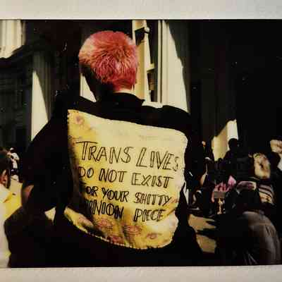 A person with pink hair wears a jacket displaying a message on the back which says "TRANS LIVES DO NOT EXIST FOR YOUR SHITTY OPINION PIECE".