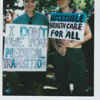 Two people are holding signs, one which says "I DON'T OWE YOU A MEDICAL TRANSITION" and the other "ACCESSIBLE HEALTHCARE FOR ALL".