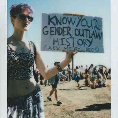 A person holds a sign reading "Know Your Gender Outlaw History" at an outdoor event.