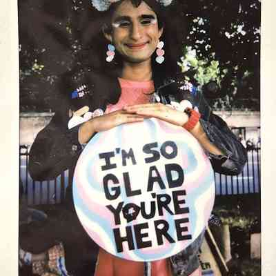 A person smiles warmly, wearing colorful pompom accessories and holding a sign that says, "I'm so glad you are here"