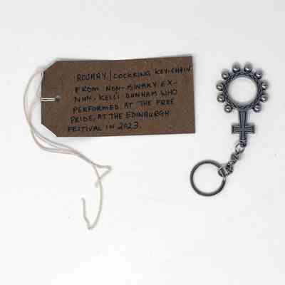 A silver rosary cock ring pendant. Next to it on the left is a brown tag with writing on it.