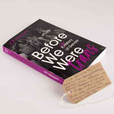 Book entitled "Before We Were trans: A NEW History of Gender" by Kit Heyam. The books spine is magenta and the cover features an old black and white photo of some formally dressed people. With a brown, handwritten luggage tag.