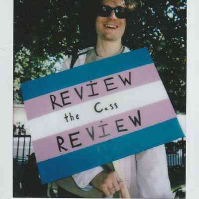 A person holding a colorful protest sign stands outside, wearing sunglasses and smiling near the trees.